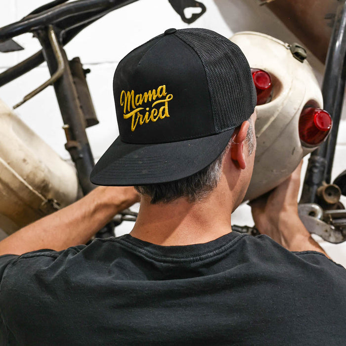 Person wearing a black cap with 'Mama Tried' in yellow text, holding a helmet.