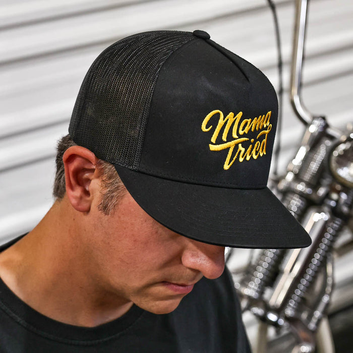 Man wearing a black cap with 'Mama Tried' in yellow text, standing next to a motorcycle.