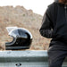 Black helmet on a guardrail with a person in dark clothing sitting nearby, against a mountainous background.